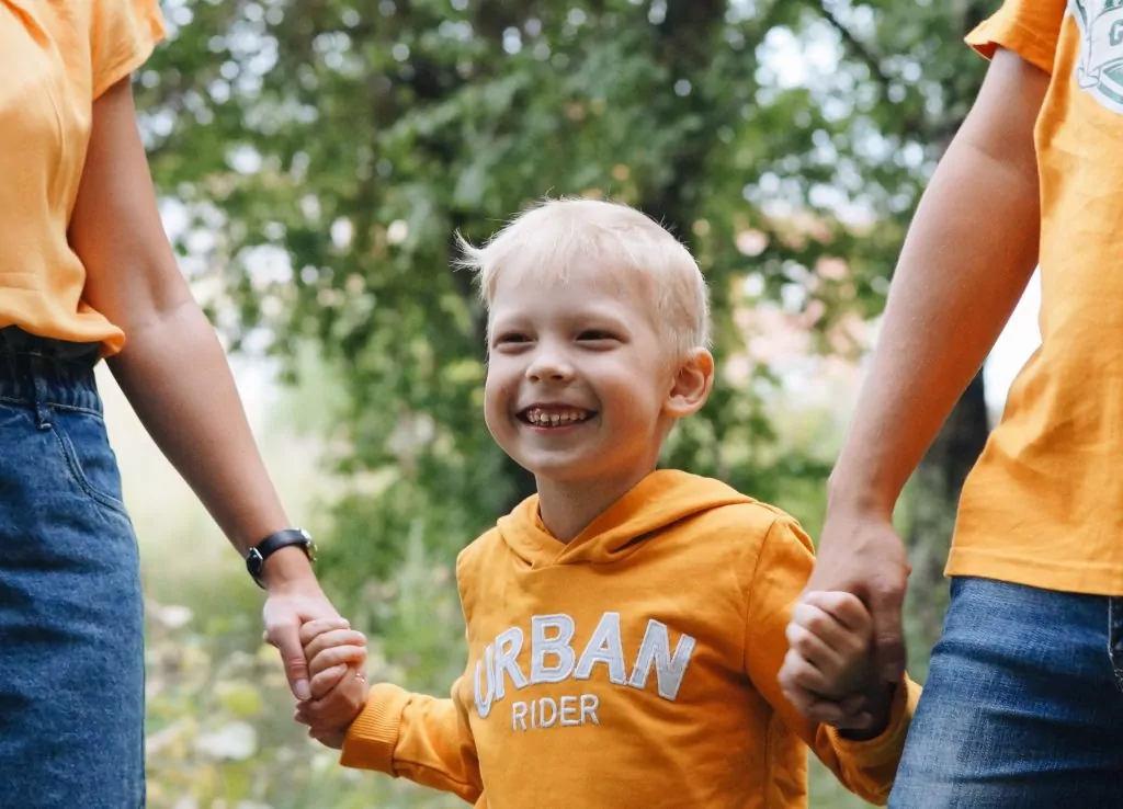 Smiling young child in an orange hoodie holds hands with two adults outdoors, standing among greenery—cherishing special Compass Family Time together.