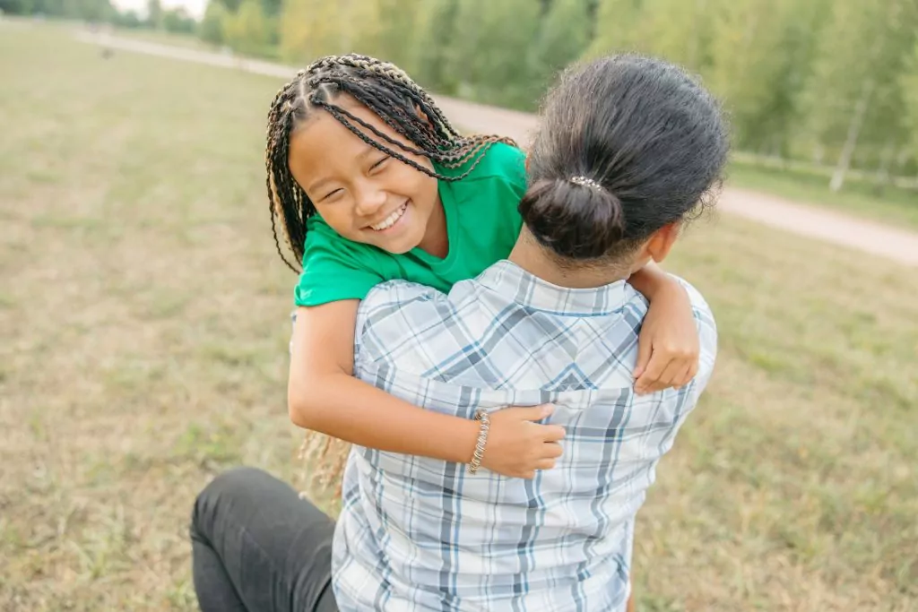 A smiling child in a green shirt hugs an adult wearing a checked shirt outdoors in a grassy area with trees in the background, capturing a joyful moment of Compass Family Time.