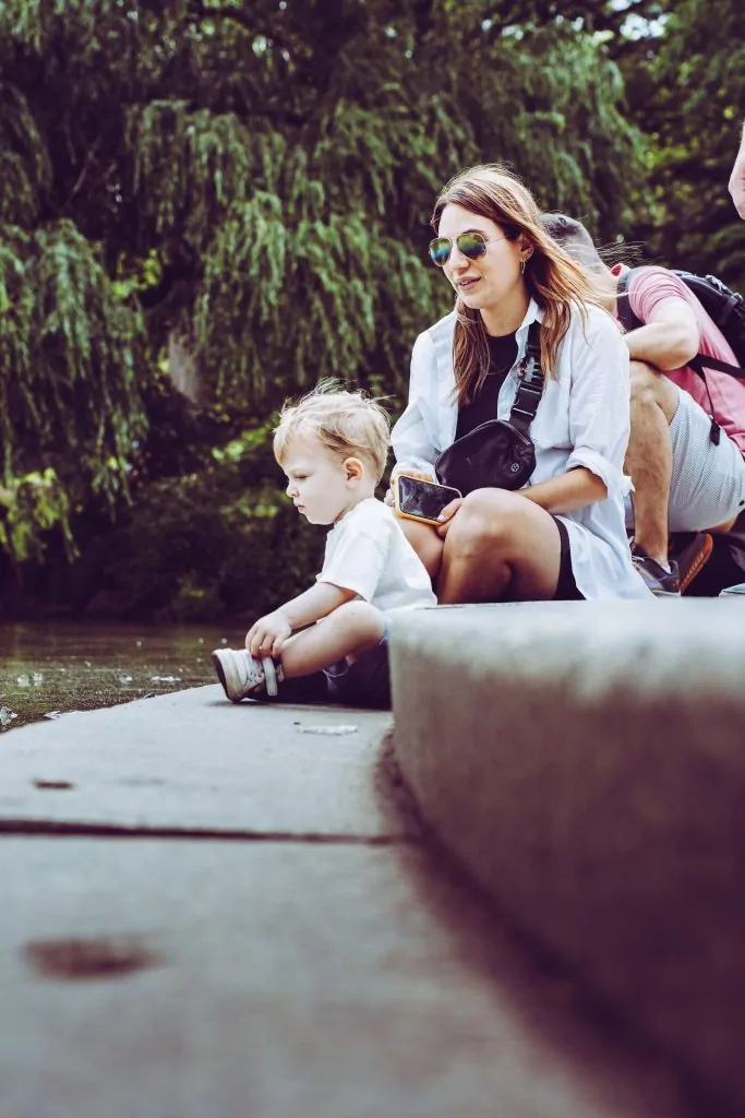 A woman and a young child sit by the edge of a pond, enjoying some Compass Family Time as they gaze at the water. Trees and other people are visible in the background.
