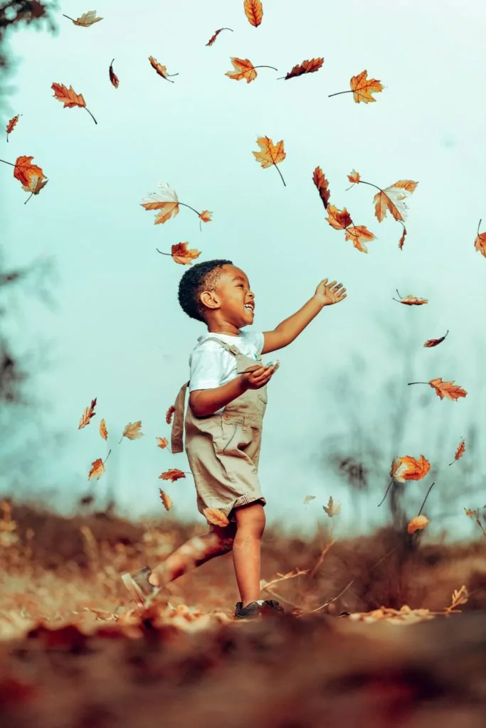 A young child in shorts and a white shirt runs outdoors, smiling with arms raised as autumn leaves fall around them on a clear day—a perfect moment of Compass Family Time.