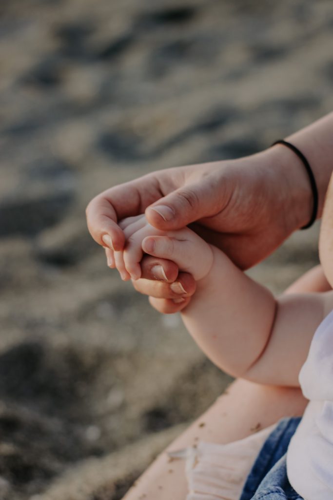 An adult hand gently holds a baby's hand, with a sandy beach backdrop visible—a perfect moment of Compass Family Time.