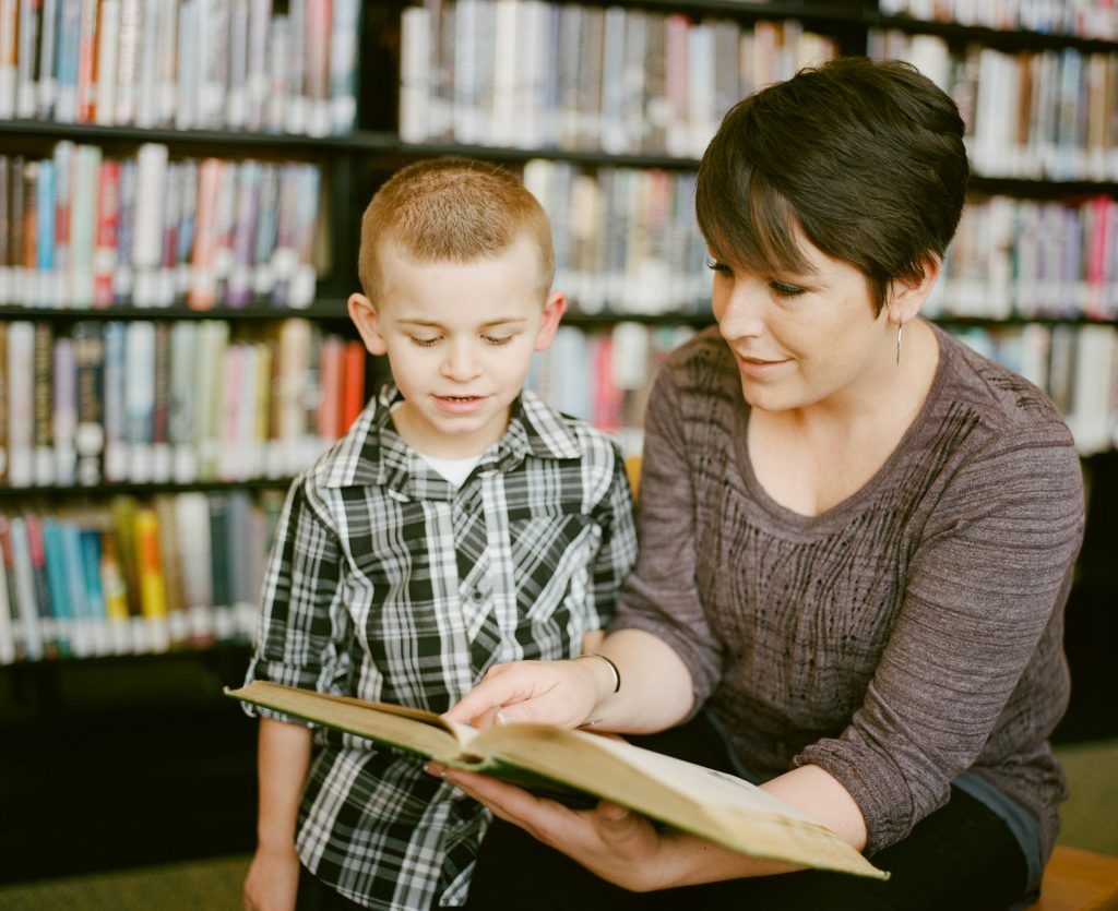 A woman sits in a library and points to a page in an open book for a young boy to read.