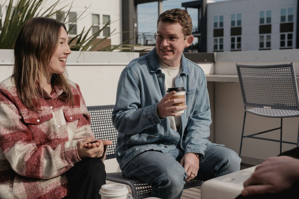 A woman and a man sitting on a rooftop terrace, talking and holding coffee cups.