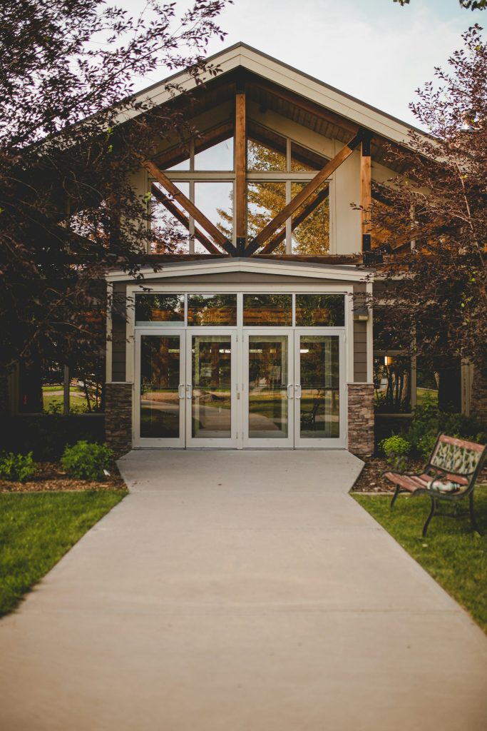 Concrete path leading to the glass-panelled entrance of a modern building with timber framing and a bench on the right.