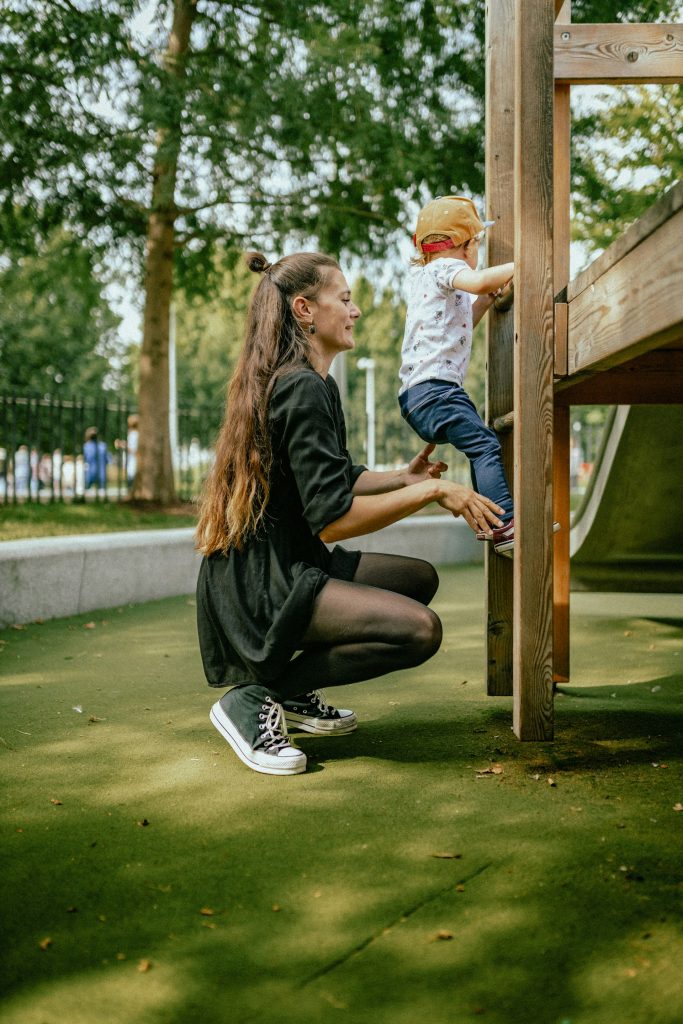 A woman in a black dress crouches to assist a toddler climbing a wooden climbing frame.