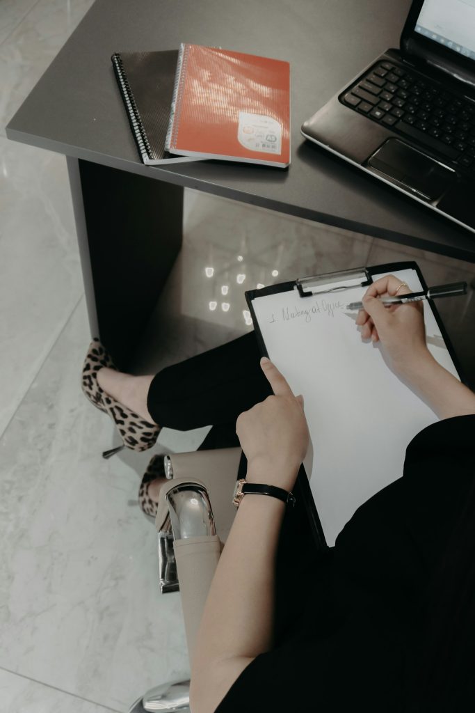 A person sitting at a desk writes “1. Meeting at Office” on a clipboard.
